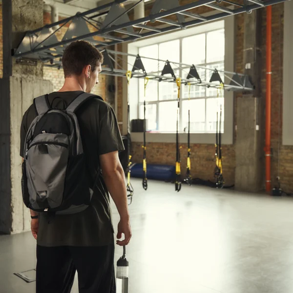 Un homme avec un sac à dos se tient dans une salle de sport, prêt à commencer un programme de fitness débutant.