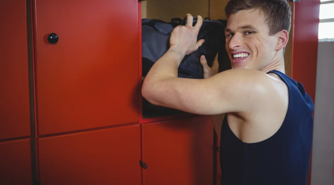 Un homme souriant tient un sac dans un casier, représentant le programme de fitness débutant.